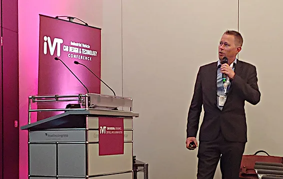 TYRI's Global R&D Manager looks sideways at a large screen on the wall whilst he holds a microphone and talks about 'the vision of future machine lighting'.