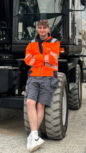 Young machine operator; Ben Wade in high-visibility workwear leaning against a heavy construction vehicle wheel, showcasing modern construction and machinery operator lifestyle