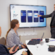 Business meeting with three colleagues discussing a TYRI mobile app interface displayed on a large screen, featuring UI designs and product prototypes on the conference table